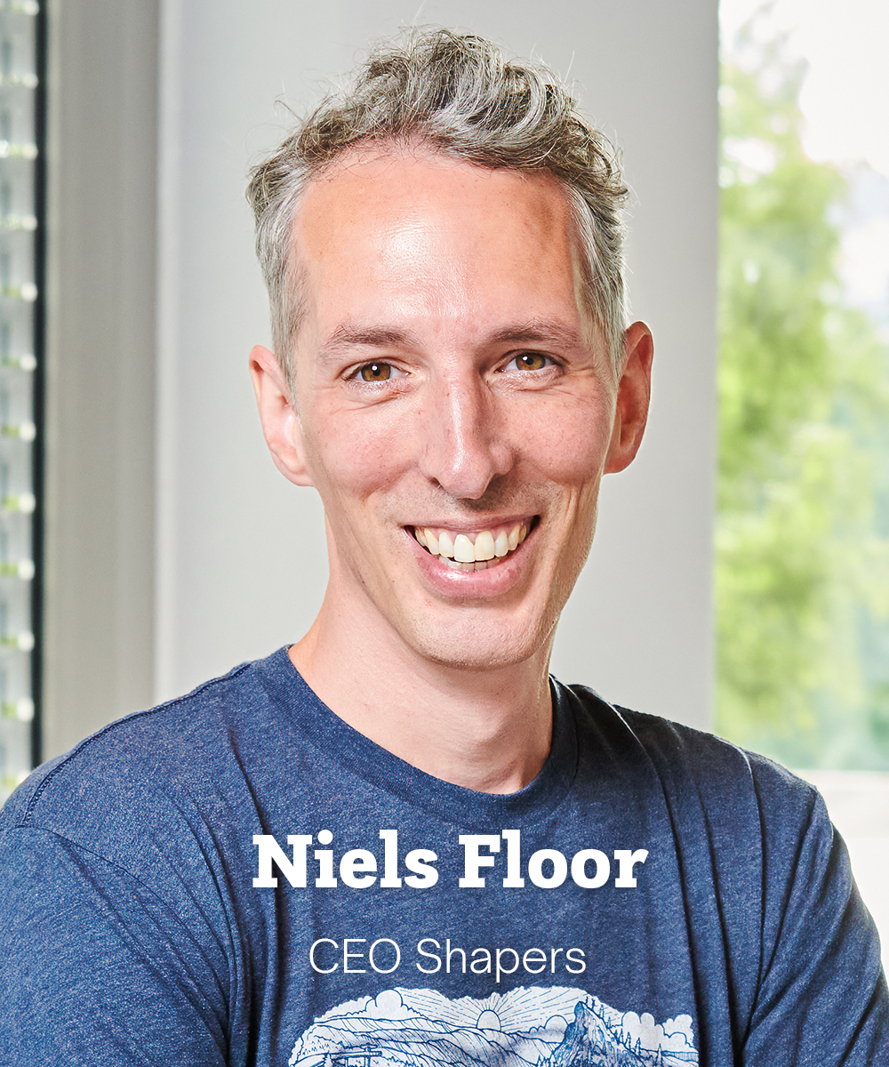 Niels has light skin and short grey curly hair. He is standing in an office smiling.