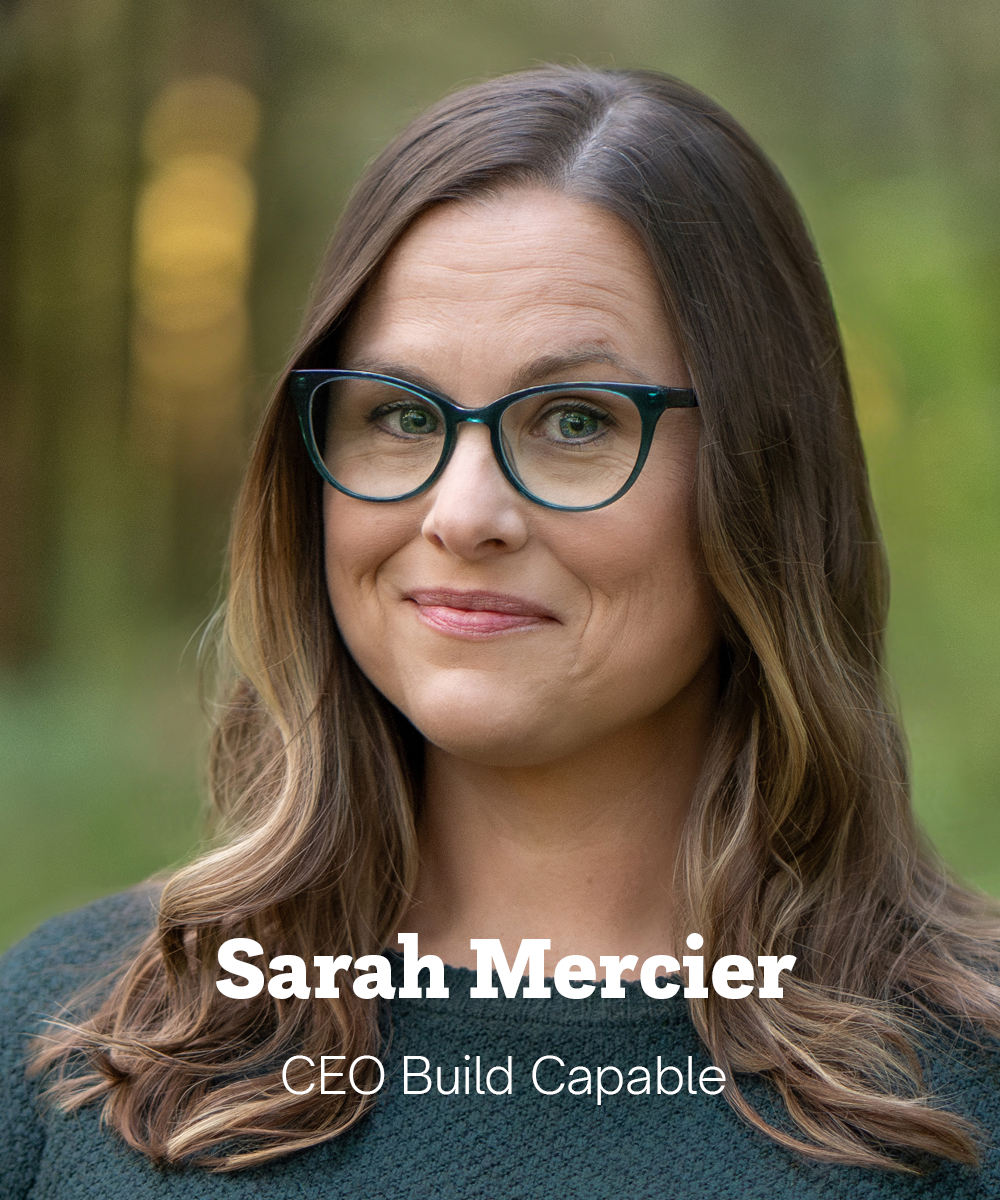 Sarah has light skin, long brown hair, and green-framed glasses. She’s standing in a forest wearing a dark green sweater and smiling slightly.