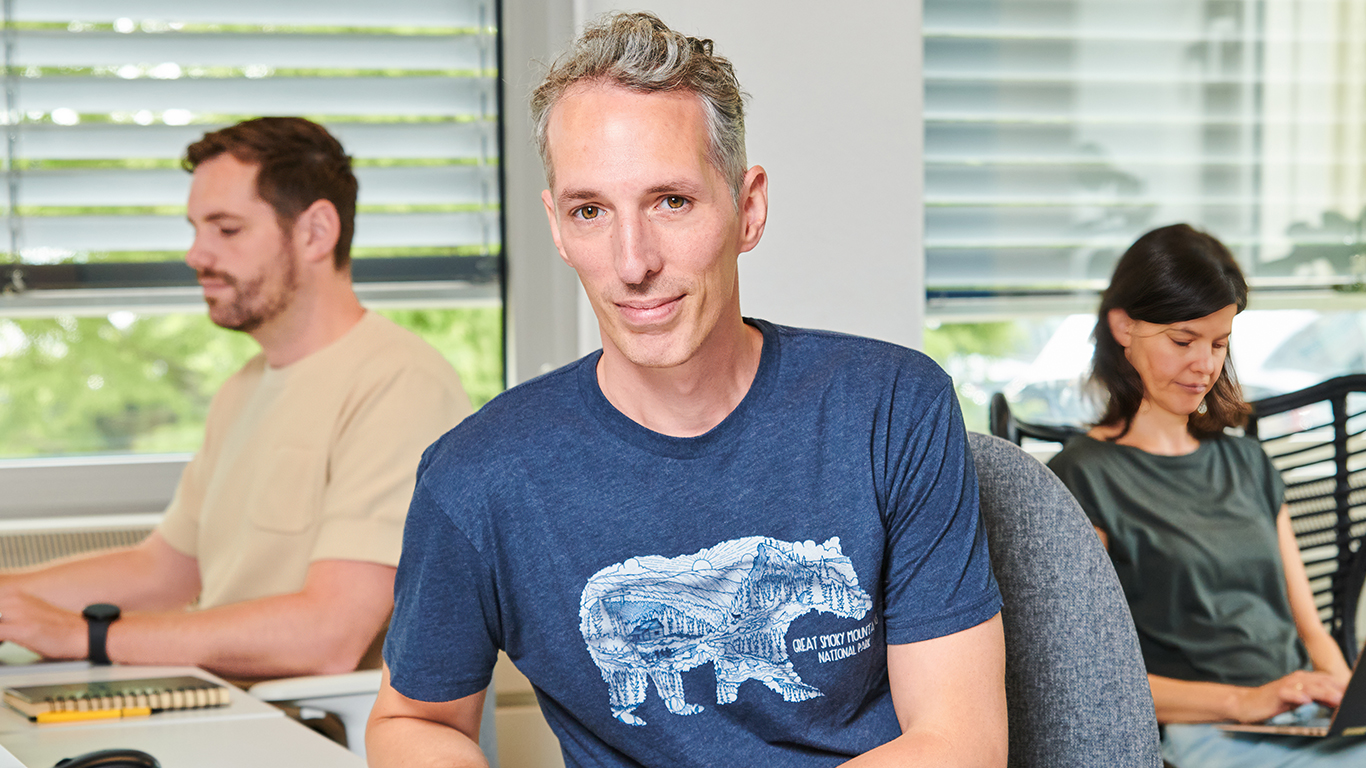 A portrait of Niels Floor, a man with short curly grey hair and light skin wearing a blue t-shirt while sitting at a desk and smiling. In the background two colleagues are working behind their computers.